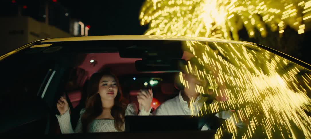 A happy young Asian couple smiles and dances while driving through a brightly lit city at night