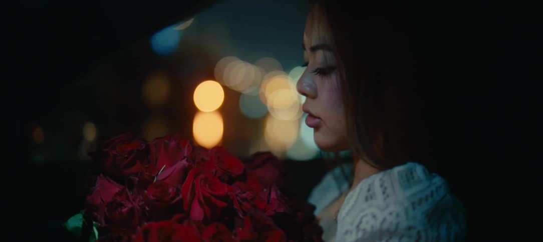 A beautiful young woman holds a bouquet of red roses while sitting in a car at night with city lights glowing in the background