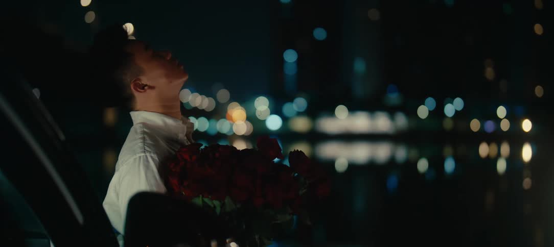A heartbroken young man smokes a cigarette while holding a bouquet of red roses, waiting alone by the water under city lights at night