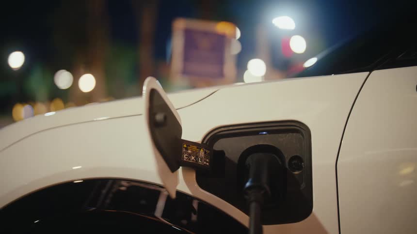 An electric car's battery recharges at a public charging station on a city street at night, with a blinking blue light indicating the process