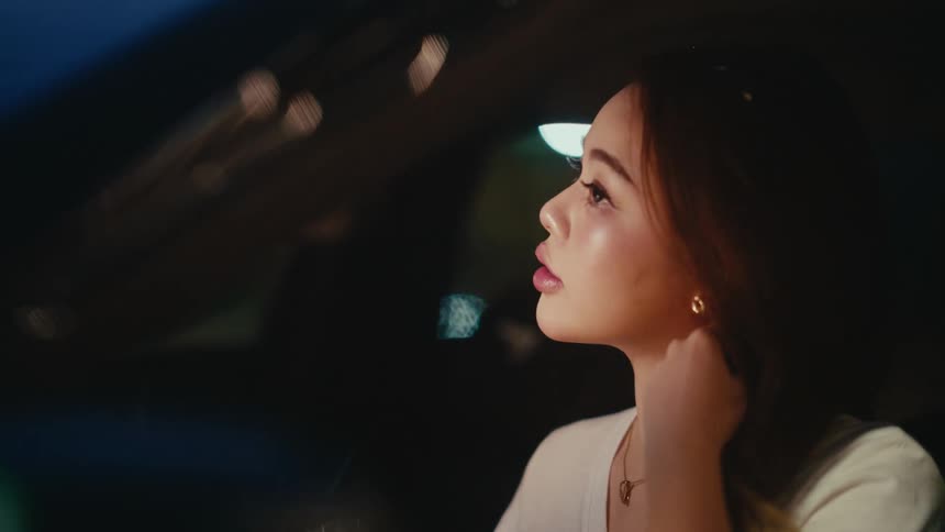 An emotional young woman sits alone in her car at night, wiping a tear from her eye in slow motion