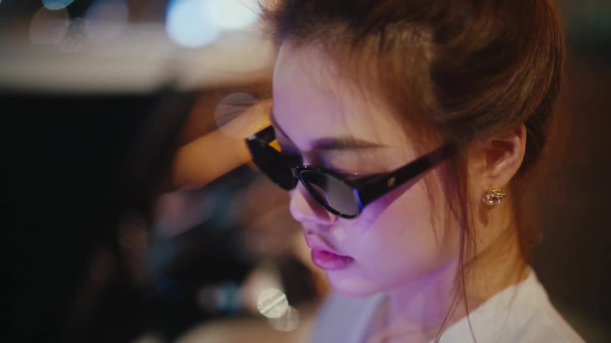 A close-up profile of a stylish young woman wearing sunglasses, her face illuminated by the vibrant city lights at night