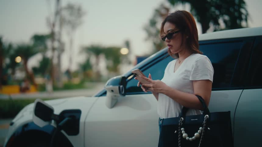 A stylish woman makes a call on her smartphone while waiting for her white electric vehicle to charge at a station