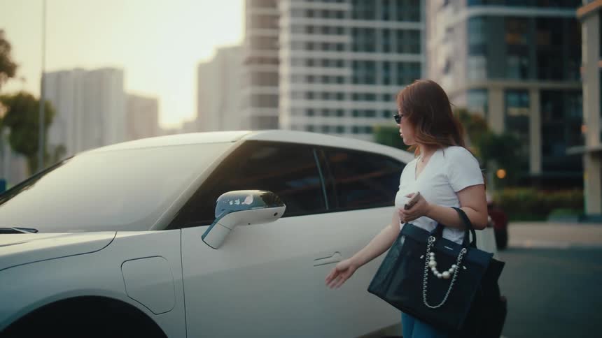 A fashionable woman exits her modern electric car, closes the door, and opens the charging port in a city at sunset