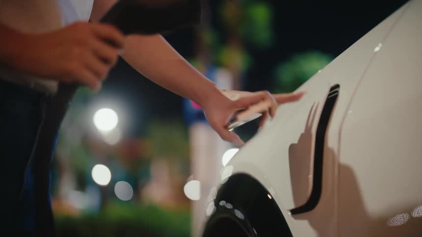 A woman uses her smartphone to unlock the charging port and plugs in the charger to her white electric vehicle at night