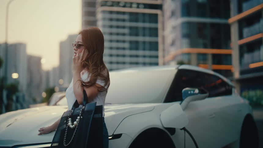 A chic businesswoman smiles while on a phone call, waiting for her luxury electric vehicle to charge in the city