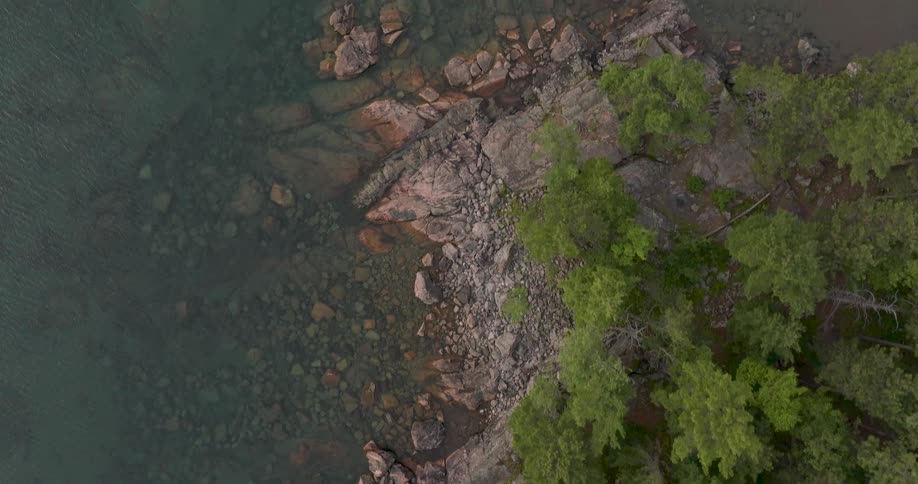 An aerial top-down shot slowly rises over a tranquil, rocky coastline revealing clear water and a lush green forest