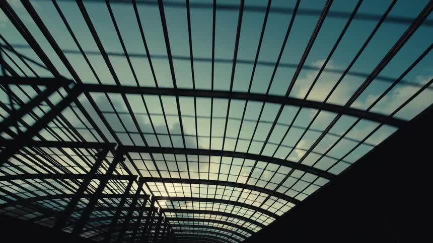 A smooth tracking shot looks up at the sky and clouds through the silhouetted framework of a modern curved glass roof