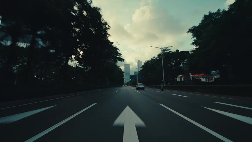 A cinematic point-of-view driving down a wide, tree-lined city road towards a modern skyline of towering skyscrapers