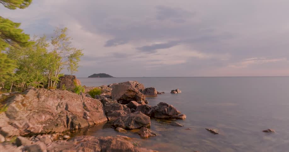 A slow aerial flyover reveals the rugged, tree-lined, rocky coastline of a vast lake during a calm and cloudy sunset