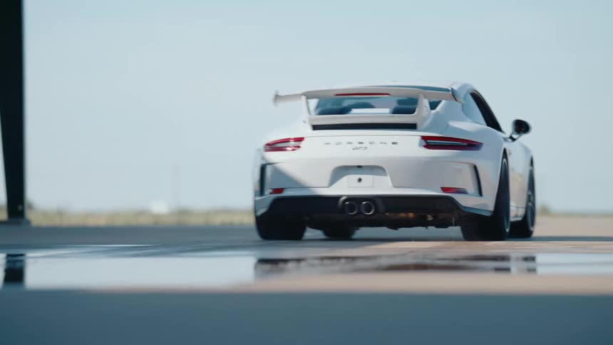 Rear view of a white Porsche GT3 sports car splashing water as it speeds away in slow motion on a bright day