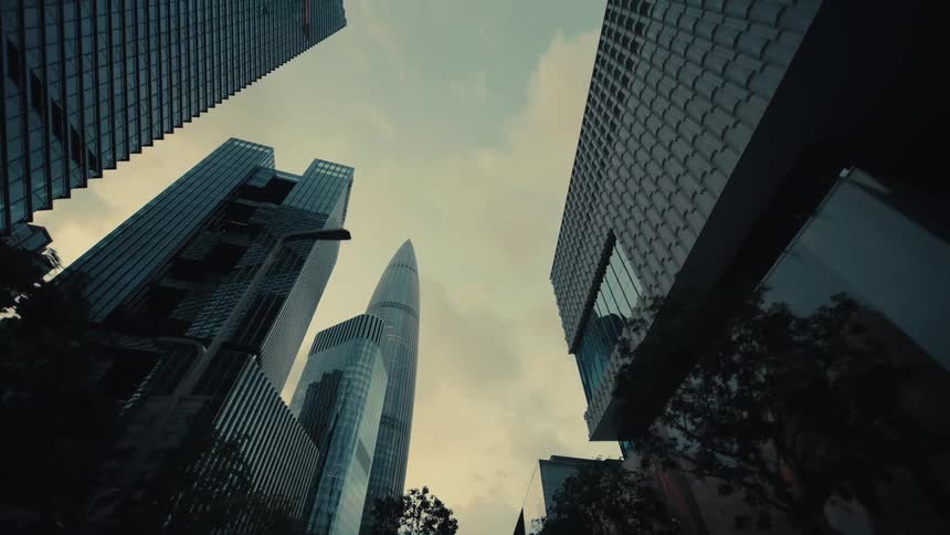 A low-angle reverse dolly shot glides past towering glass skyscrapers in a futuristic city at dusk