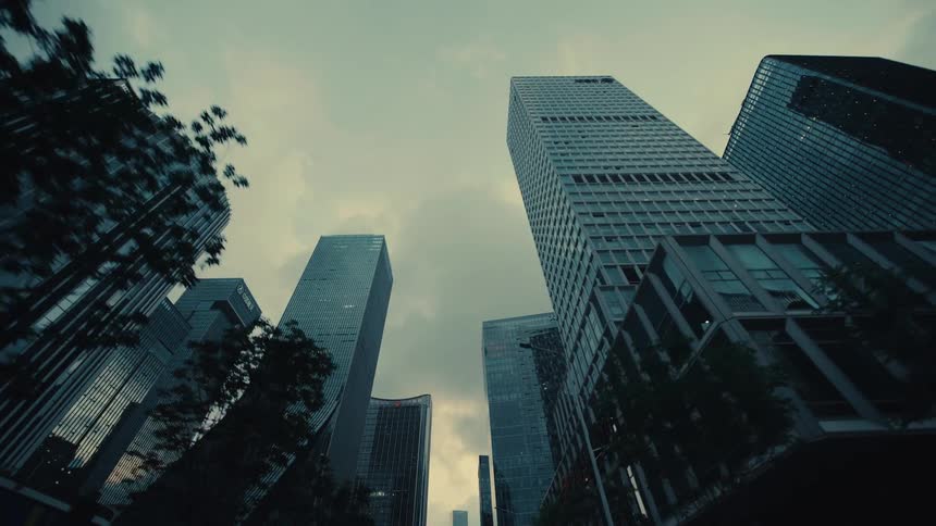 A moody, low-angle tracking shot looking up while driving through a modern financial district with towering glass skyscrapers