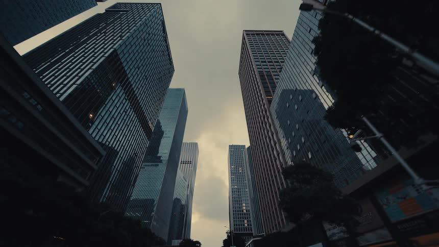 A slow, low-angle tracking shot through a modern city canyon of towering glass skyscrapers reaching for the overcast sky