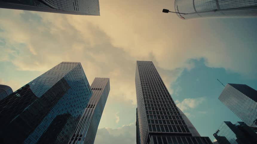 A slow, low-angle tracking shot through a modern city's financial district reveals towering glass skyscrapers against a dramatic sky