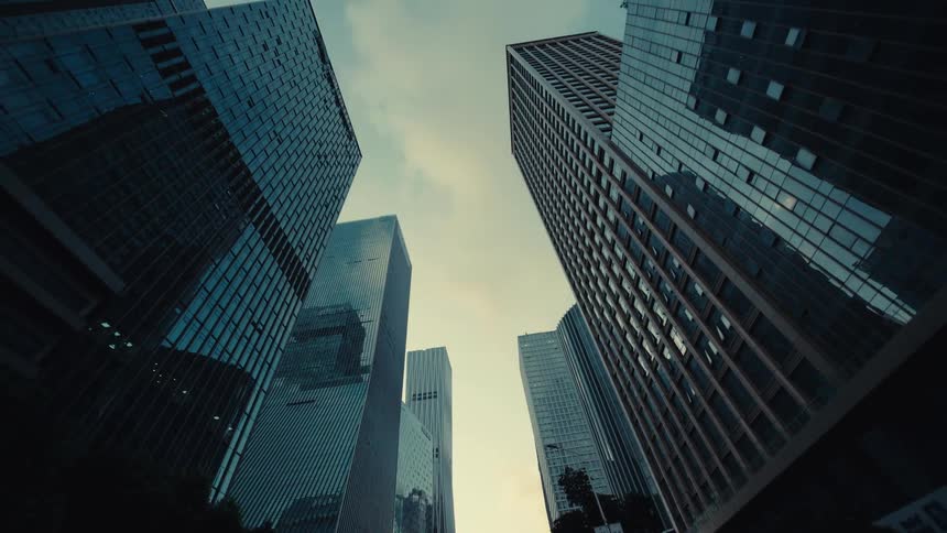A smooth, low-angle tracking shot moves backward along a city street, looking up at towering modern glass skyscrapers against the sky