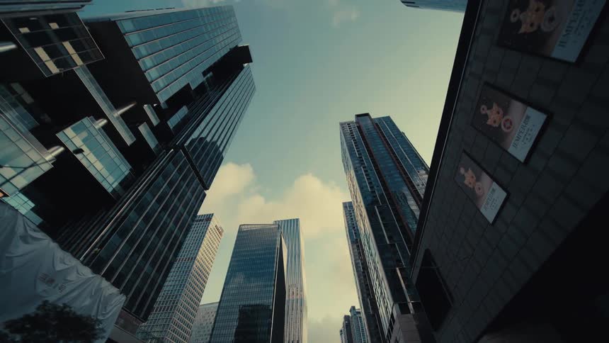 An impressive low-angle tracking shot moves backward through a modern financial district, revealing towering glass skyscrapers against a blue sky