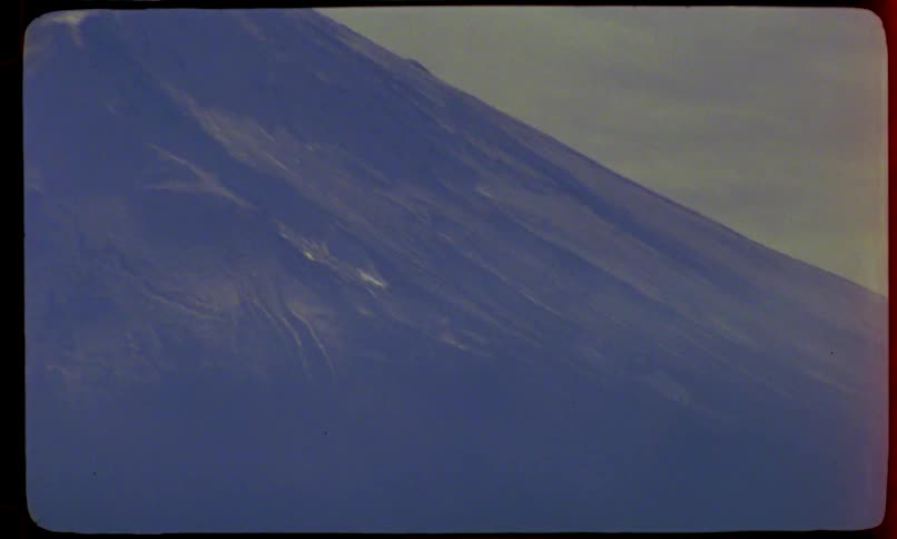 A vintage, archival shot pans down from the peak of Mount Fuji, revealing its massive slope and the surrounding landscape