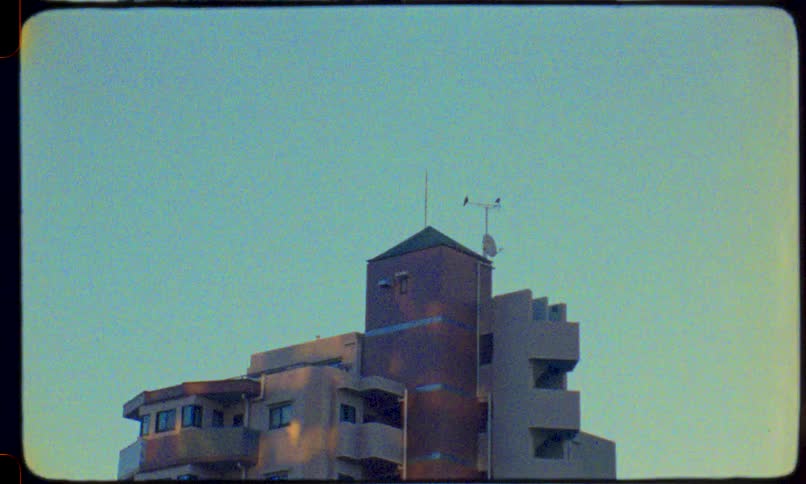 Vintage 8mm film captures a static shot of a city building with a weather vane spinning in the wind