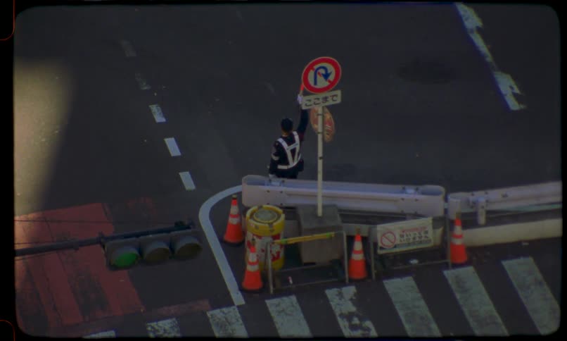 Vintage 8mm-style footage from a high angle shows a traffic warden directing a yellow taxi cab at a city intersection in Japan