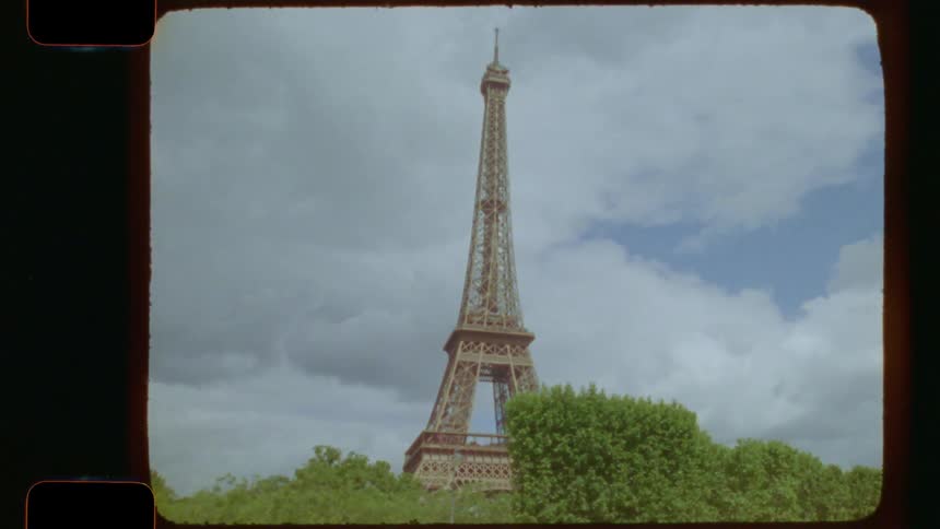 A vintage film tracking shot reveals the iconic Eiffel Tower in Paris from behind lush green trees on a cloudy day