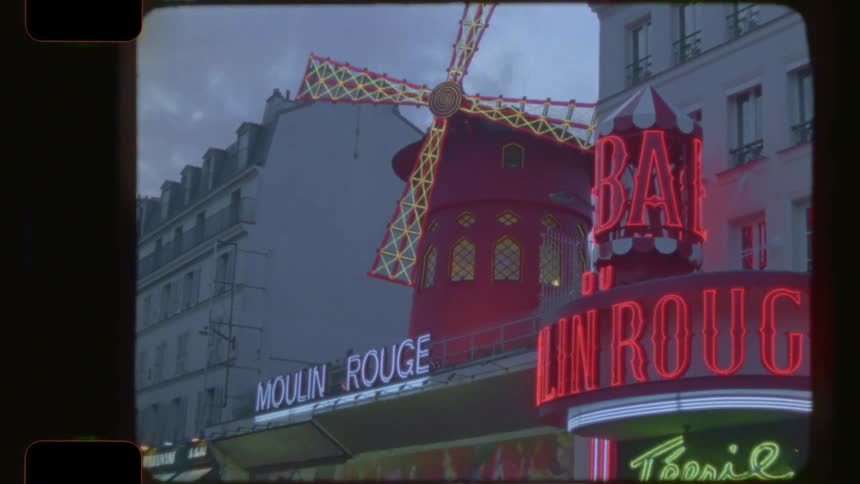 Vintage 16mm film footage zooms out from the iconic windmill to reveal the neon signs of the Moulin Rouge in Paris at dusk