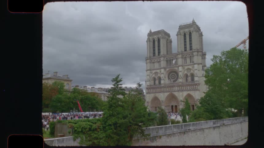 Vintage 16mm film captures a static wide shot of the Notre-Dame Cathedral in Paris on a cloudy day with crowds of tourists gathered