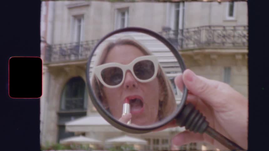 Vintage 8mm footage shows a stylish blonde woman in sunglasses applying red lipstick in her scooter's rearview mirror on a Parisian street