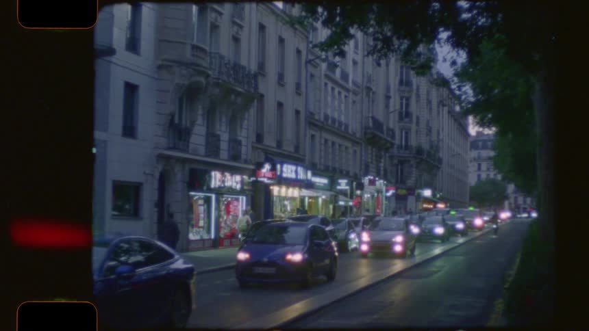 Vintage 16mm film captures a bustling Paris street at dusk with a long line of taxi traffic and illuminated storefronts