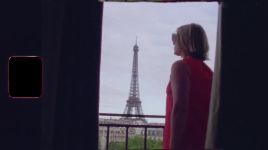 A stylish woman in a red dress puts on sunglasses and looks out over the Eiffel Tower from her Parisian balcony in this vintage film footage