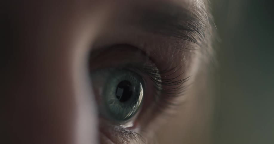Extreme close-up of a human eye with a screen reflected in the pupil as it blinks in a dimly lit room