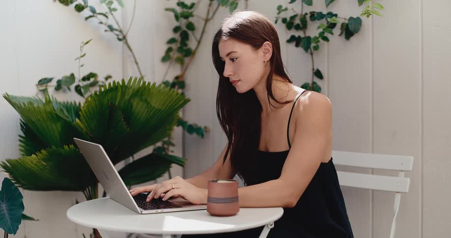 A stylish young woman works remotely on her laptop while sitting at a table on a sunlit, plant-filled patio