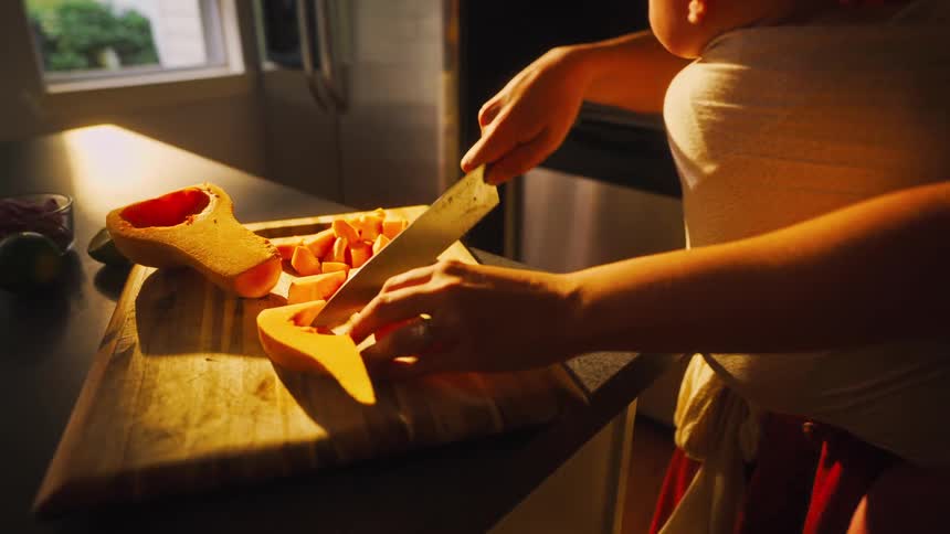 Close up of a mother holding her baby in a sling while chopping butternut squash in a warm, sunlit kitchen