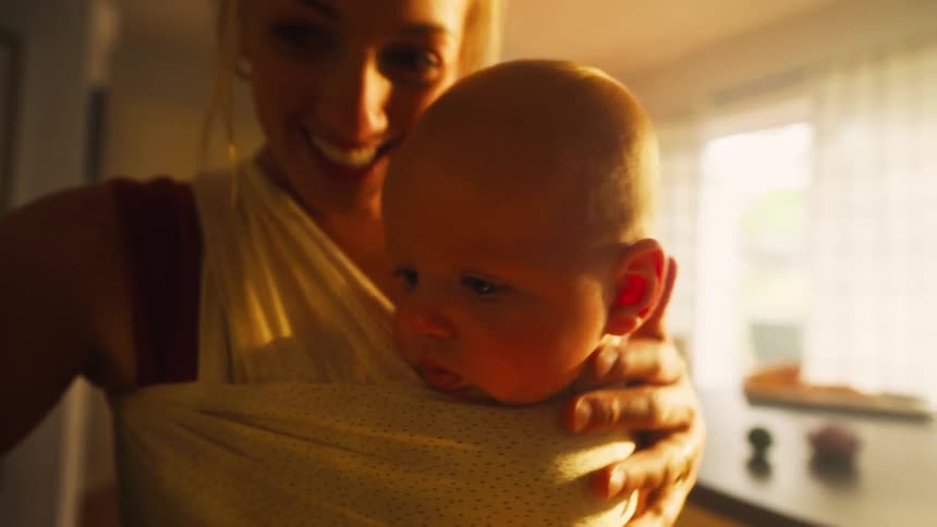 Close-up of a content newborn baby nestled in a sling against its smiling mother, illuminated by warm evening light