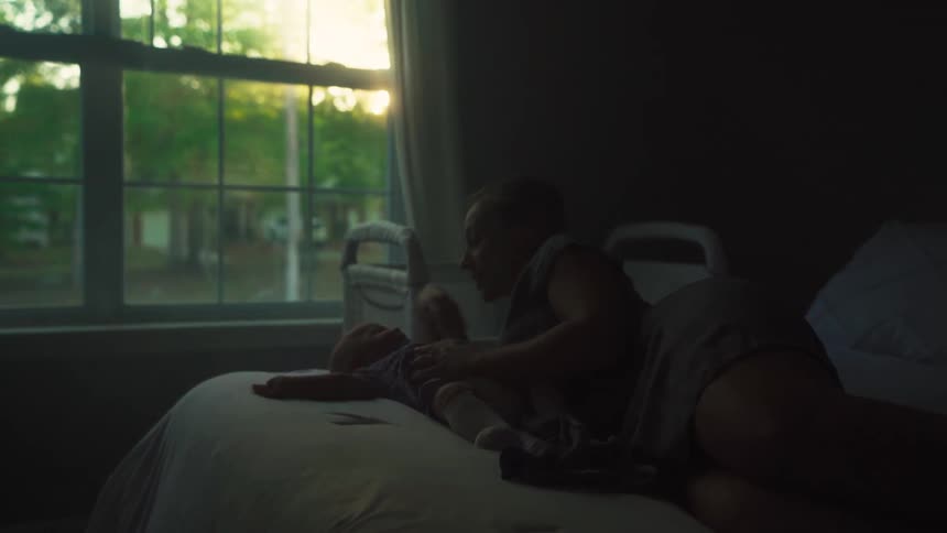 A mother and her newborn baby share an intimate moment on a bed, silhouetted by the warm light of the sun from a nearby window