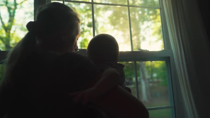 A young mother and her newborn baby are silhouetted as they look out a window into the golden hour sunlight