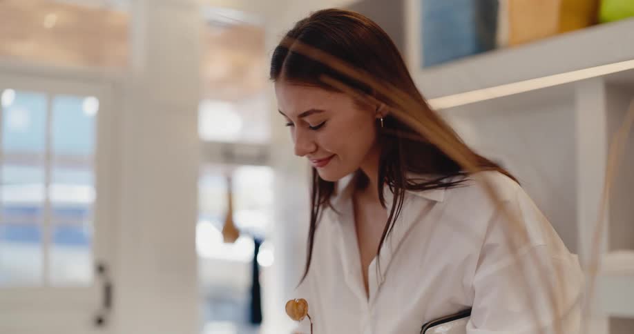 A stylish young woman in a white shirt smiles happily as she browses a bright, modern clothing boutique in slow motion
