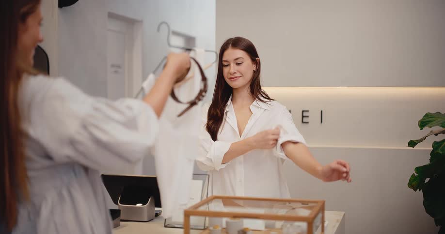 A friendly female shop assistant smiles and prepares to serve a customer at the counter of a stylish clothing boutique