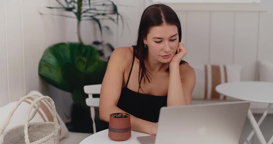 A focused young woman rests her head in her hand while intently working on her laptop in a bright, modern space