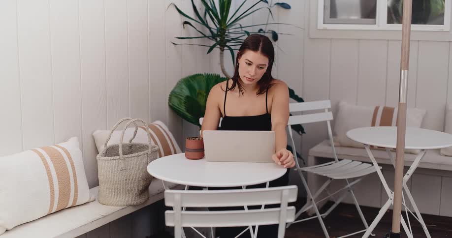 A focused young woman works remotely on her laptop while sitting at a bright, modern outdoor patio
