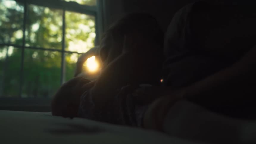 Silhouetted mother and baby share a tender moment on a bed, backlit by the warm glow of the setting sun