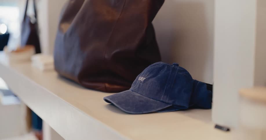 Slow push-in on a blue denim 'SURF' cap and a brown leather tote bag displayed on a minimalist boutique shelf