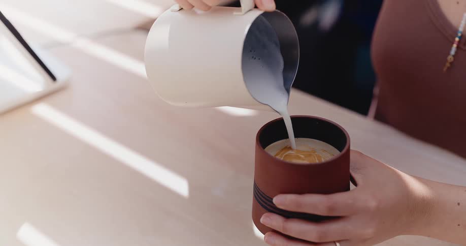 A close-up slow-motion shot of a person pouring frothed milk from a pitcher into a stylish brown ceramic cup of espresso to make a latte