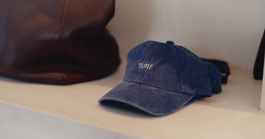 A close-up of a blue baseball cap with the word 'SURF' embroidered on it, resting on a white shelf next to a leather bag