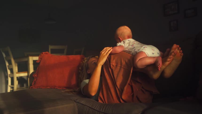 A mother playfully lifts her newborn baby with her legs while lying on the couch in a warm, sunlit room