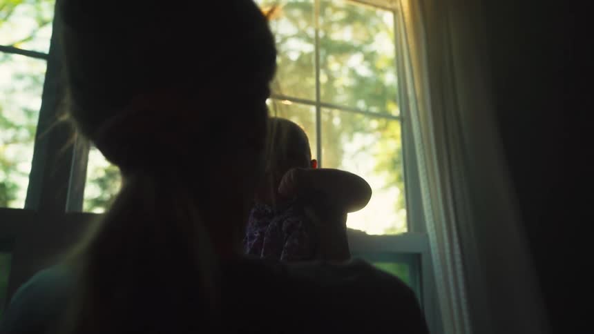 A tender moment as a silhouetted mother holds and comforts her newborn baby in front of a sunlit window