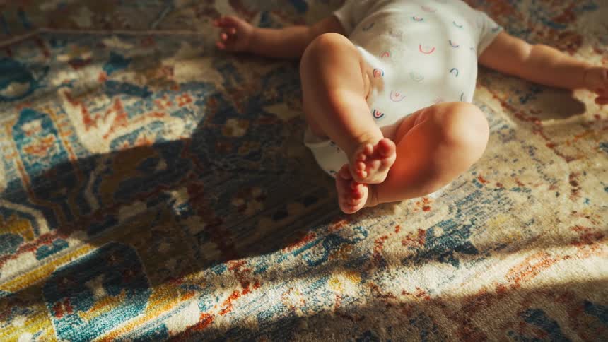 A newborn baby rests on a colorful patterned rug with its tiny feet crossed as sunlight streams into the room