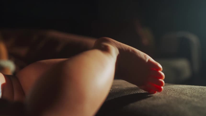A close-up shot of a baby's tiny leg and foot as they lie on a couch in a warm patch of sunlight