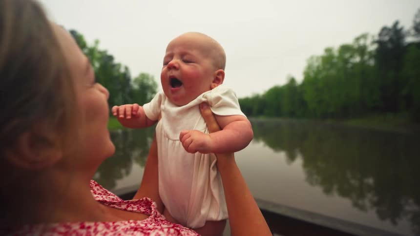 A young mother affectionately holds and kisses her yawning newborn baby outdoors by a serene lake