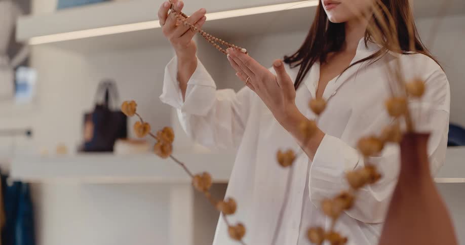A stylish young woman in a white shirt carefully examines a beaded necklace in a modern, minimalist boutique